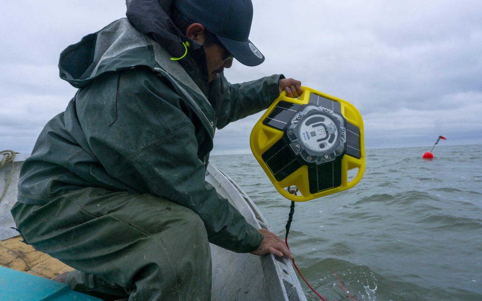 A person in rain gear leans over the side of a skiff to lower a yellow Sofar Spotter buoy into Kuskokwim Bay, with a red marker buoy floating in the distance.