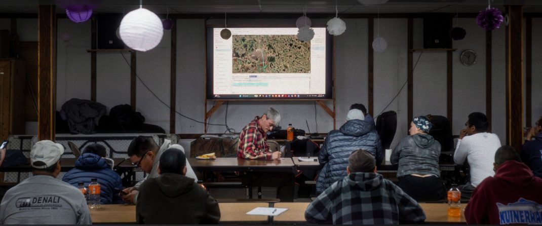 A full room of trainees seated at long tables watching a projected mapping session in a Quinhagak community hall.