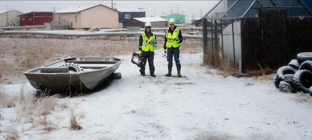 Two volunteers in high-visibility vests carry a drone case across snow-covered ground in Quinhagak, with village houses and a skiff behind them.