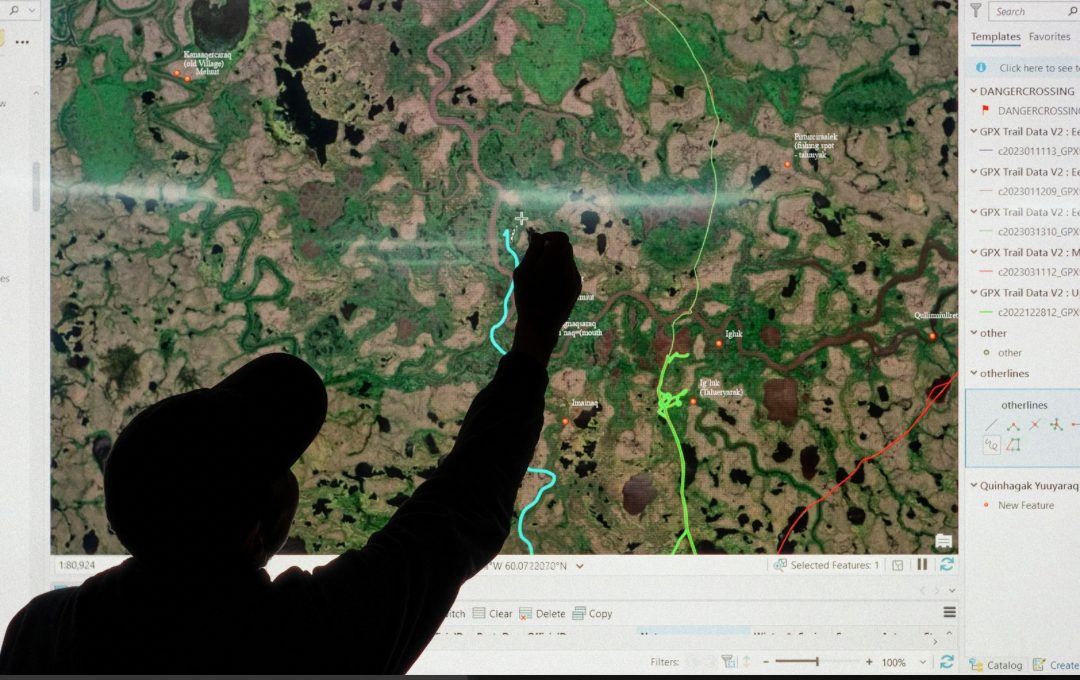 A person in silhouette reaches up to a projected satellite map of the Kanektok River area, tracing a river channel among labelled Yup'ik place names.