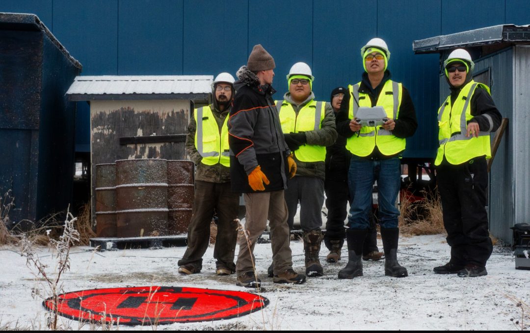Six Quinhagak Search and Rescue members in high-visibility vests and hard hats stand in the snow around a drone controller, with a landing pad in the foreground.