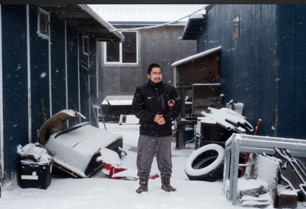 Cik Mark stands in falling snow in a yard between two blue buildings, among a snow-covered sled, tires and stacked gear.